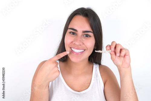 Young beautiful brunette woman wearing white top over white wall holding an invisible aligner and pointing to her perfect straight teeth. Dental healthcare and confidence concept.