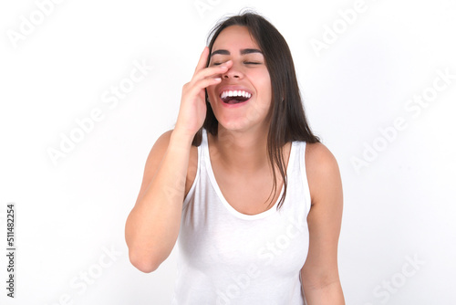 young beautiful brunette woman wearing white top over white wall makes face palm and smiles broadly, giggles positively hears funny joke poses
