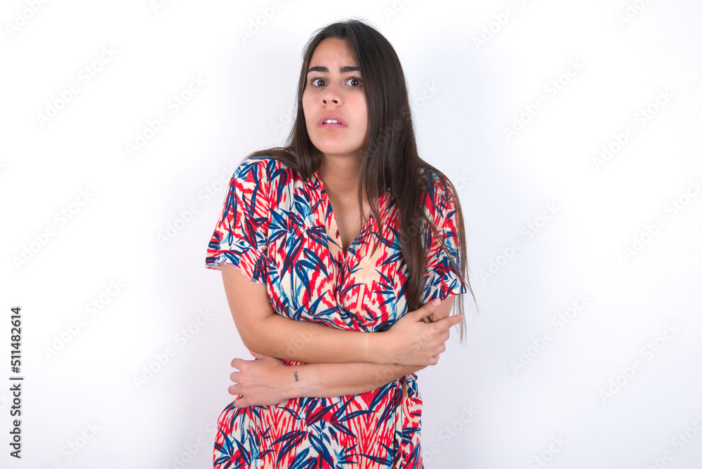 young beautiful brunette woman wearing colourful dress over white wall shaking and freezing for winter cold with sad and shock expression on face. young beautiful brunette woman wearing colourful dress over white wall shaking and freezing for winter cold with sad and shock expression on face.