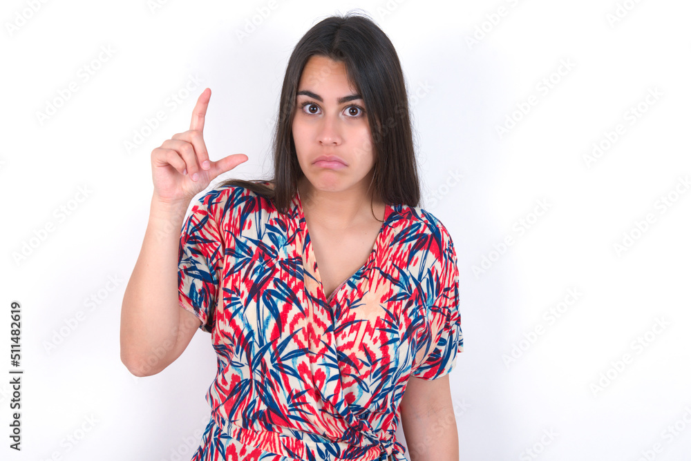 young beautiful brunette woman wearing colourful dress over white wall purses lip and gestures with hand, shows something very little. young beautiful brunette woman wearing colourful dress over white wall purses lip and gestures with hand, shows something very little.
