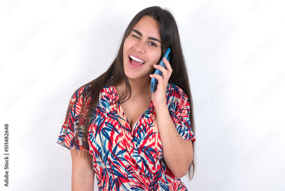 Funny young beautiful brunette woman wearing colourful dress over white wall laughs happily, has phone conversation, being amused by friend, closes eyes. Funny young beautiful brunette woman wearing colourful dress over white wall laughs happily, has phone conversation, being amused by friend, closes eyes.