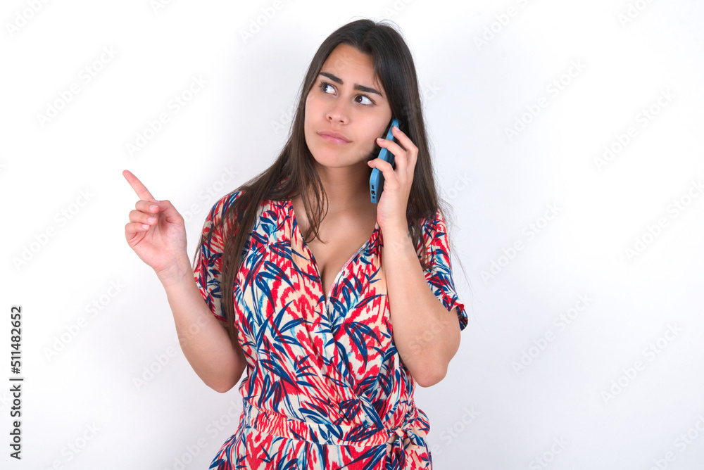 young beautiful brunette woman wearing colourful dress over white wall speaks on mobile phone spends free time indoors calls to friend.