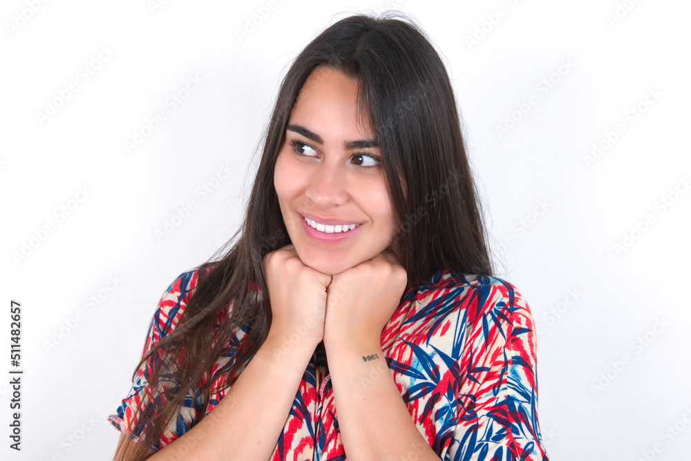 young beautiful brunette woman wearing colourful dress over white wall holds hands under chin, glad to hear heartwarming words from stranger young beautiful brunette woman wearing colourful dress over white wall holds hands under chin, glad to hear heartwarming words from stranger