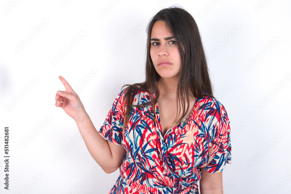 Serious young beautiful brunette woman wearing colourful dress over white wall smirks face points away on copy space shows something unpleasant. Look at this advertisement. Big price concept Serious young beautiful brunette woman wearing colourful dress over white wall smirks face points away on copy space shows something unpleasant. Look at this advertisement. Big price concept
