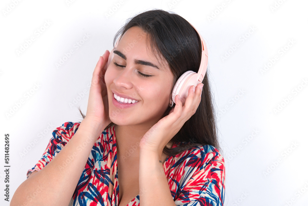 Pleased young beautiful brunette woman wearing colourful dress over white wall enjoys listening pleasant melody keeps hands on stereo headphones closes eyes. Spending free time with music Pleased young beautiful brunette woman wearing colourful dress over white wall enjoys listening pleasant melody keeps hands on stereo headphones closes eyes. Spending free time with music