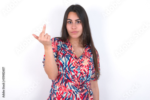 young beautiful brunette woman wearing colourful dress over white wall shows middle finger bad sign asks not to bother. Provocation and rude attitude. photo
