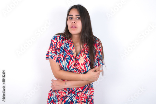 young beautiful brunette woman wearing colourful dress over white wall shaking and freezing for winter cold with sad and shock expression on face.