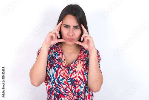 Serious concentrated young beautiful brunette woman wearing colourful dress over white keeps fingers on temples, tries to ease tension, gather with thoughts and remember important information for exam