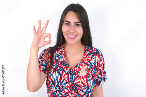young beautiful brunette woman wearing colourful dress over white wall hold hand arm okey symbol toothy approve advising novelty news