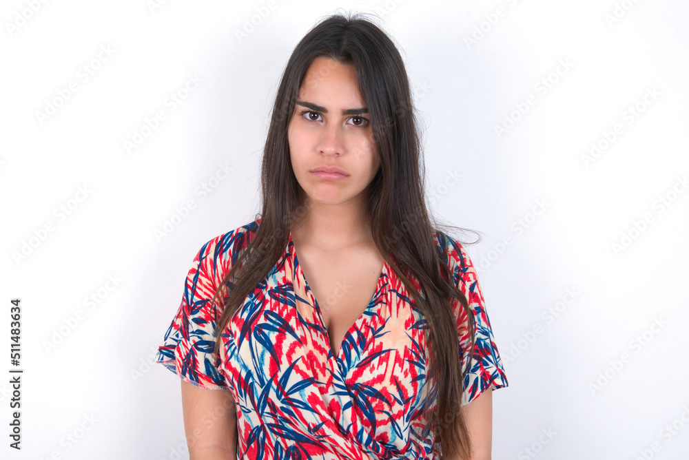 Displeased young beautiful brunette woman wearing colourful dress over white wall frowns face feels unhappy has some problems. Negative emotions and feelings concept Displeased young beautiful brunette woman wearing colourful dress over white wall frowns face feels unhappy has some problems. Negative emotions and feelings concept