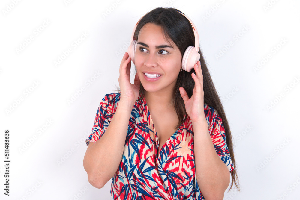 young beautiful brunette woman wearing colourful dress over white wall wears stereo headphones listens music concentrated aside. People hobby lifestyle concept young beautiful brunette woman wearing colourful dress over white wall wears stereo headphones listens music concentrated aside. People hobby lifestyle concept