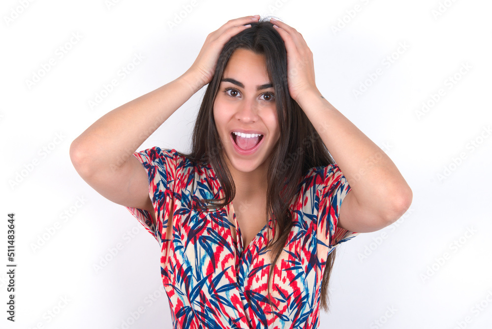 Cheerful overjoyed young beautiful brunette woman wearing colourful dress over white wall reacts rising hands over head after receiving great news. Cheerful overjoyed young beautiful brunette woman wearing colourful dress over white wall reacts rising hands over head after receiving great news.