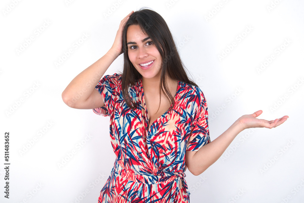 Shocked amazed surprised young beautiful brunette woman wearing colourful dress over white wall hold hand offering proposition Shocked amazed surprised young beautiful brunette woman wearing colourful dress over white wall hold hand offering proposition
