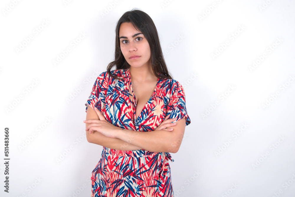 Confident young beautiful brunette woman wearing colourful dress over white wall with arms crossed looking to the camera Confident young beautiful brunette woman wearing colourful dress over white wall with arms crossed looking to the camera