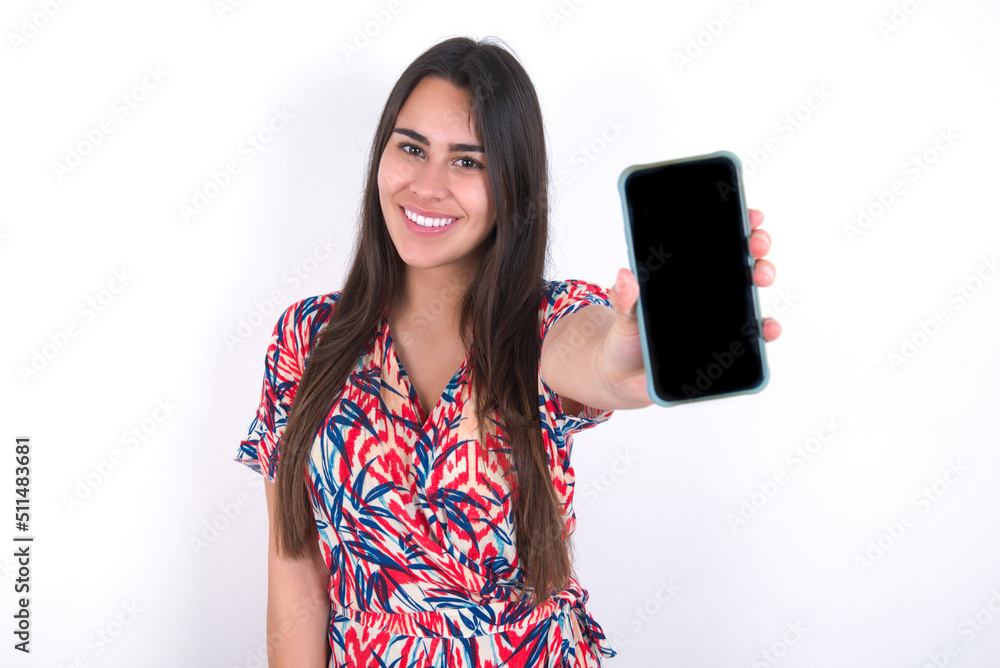 Charming adorable young beautiful brunette woman wearing colourful dress over white wall holding modern device, showing black screen smartphone