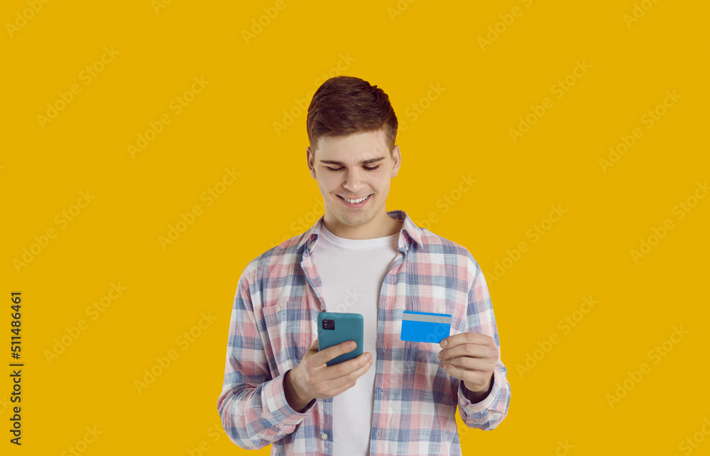 Happy guy with mobile phone and credit card makes online purchases using bank's mobile application. Young man with smartphone and bank card isolated on orange background. Online payment concept. Happy guy with mobile phone and credit card makes online purchases using bank's mobile application. Young man with smartphone and bank card isolated on orange background. Online payment concept.