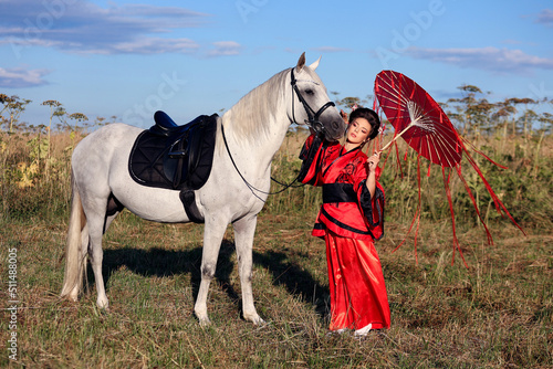 Beautifil dark haired girl in traditional Japanese dress with red umbrella standing with white horse in the field