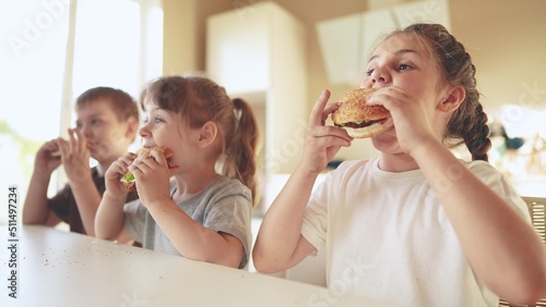children eat burgers. fast food burger. a group fun of small children in the kitchen greedily eat fast food burgers. big family small kids having breakfast in the morning in the kitchen eating burger