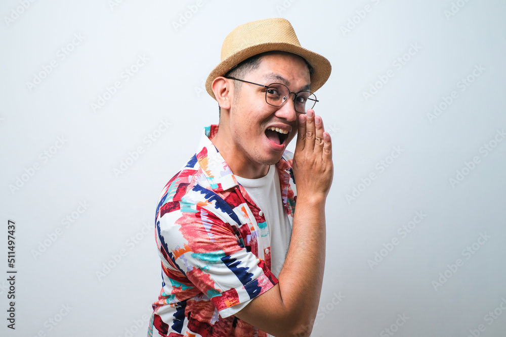 Portrait of happy Asian tourist in casual shirt, bucket hat and trendy sunglasses standing and doing whisper gesture with han. Traveling, holiday trip, summer vacation concept Portrait of happy Asian tourist in casual shirt, bucket hat and trendy sunglasses standing and doing whisper gesture with han. Traveling, holiday trip, summer vacation concept