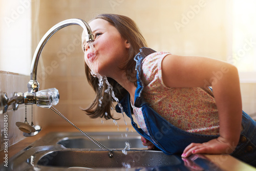 The thirst is real. Shot of a little girl drinking water directly from the kitchen tap at home. photo