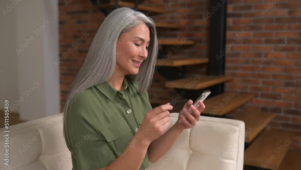 Middle-aged Asian woman consumer holding credit card and smartphone sitting on sofa at home making online banking transaction. Multiracial gray-haired lady using mobile for shopping online Middle-aged Asian woman consumer holding credit card and smartphone sitting on sofa at home making online banking transaction. Multiracial gray-haired lady using mobile for shopping online