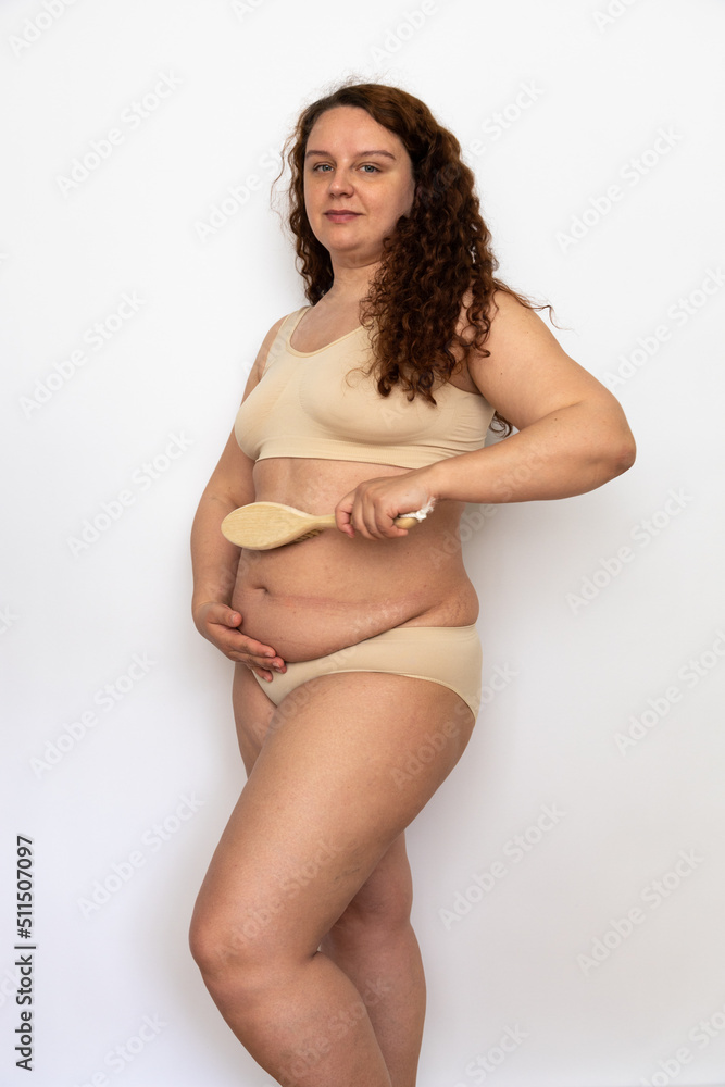 Side view of overweight middle-aged woman with curly hair wearing beige underwear, rubbing excess belly with wooden brush, posing on white background. Weight loss, lymphatic drainage, anti-cellulite. Side view of overweight middle-aged woman with curly hair wearing beige underwear, rubbing excess belly with wooden brush, posing on white background. Weight loss, lymphatic drainage, anti-cellulite.