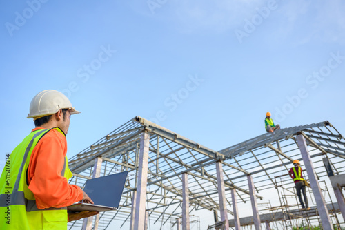 male construction engineer holding laptop standing at a house construction site Controlling and ordering Construction workers for quality inspection and steel roof truss design plans