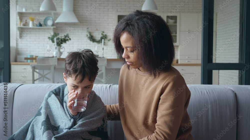 A worried and sad young mother gently wraps her sick son with a warm blanket and gives him a glass of water to drink. Boy with signs of infectious disease and fever leaning on caring mother. A worried and sad young mother gently wraps her sick son with a warm blanket and gives him a glass of water to drink. Boy with signs of infectious disease and fever leaning on caring mother.