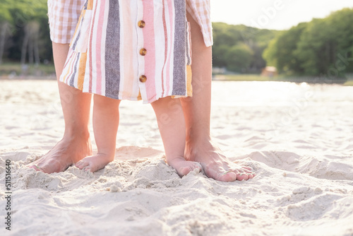 close up legs, mom teaches daughter to walk holding hands on beach on sand on sunny day