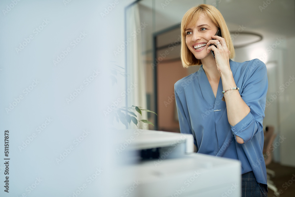 Happy businesswoman making phone call while using photocopier in office. Happy businesswoman making phone call while using photocopier in office.