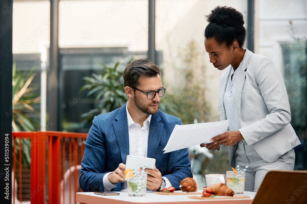 Mid adult businessman using touchpad while analyzing paperwork with African American colleague in cafe. Mid adult businessman using touchpad while analyzing paperwork with African American colleague in cafe.