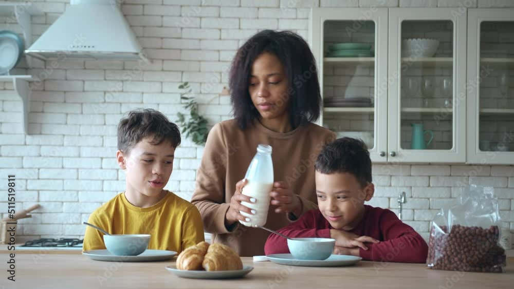 Little boys sit at kitchen table, waiting for breakfast. Young mother pours milk into bowls and sprinkles cereal chocolate flakes for her sons , preparing delicious healthy breakfast in the morning Little boys sit at kitchen table, waiting for breakfast. Young mother pours milk into bowls and sprinkles cereal chocolate flakes for her sons , preparing delicious healthy breakfast in the morning