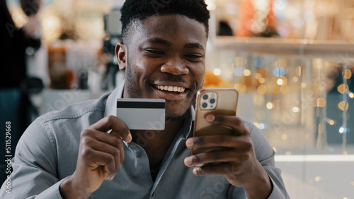 Satisfied african american man makes purchase in online store pays for goods with credit card on mobile phone uses e-banking application happy young male making safe transaction secure payment concept