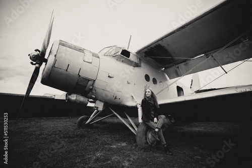 Fototapeta Naklejka Na Ścianę i Meble -  Art work. A beautiful girl is sitting on the wing of an airplane, she is bored. Getting ready to fly. The plane stands on the grass in a clearing. The photograph is black and white.