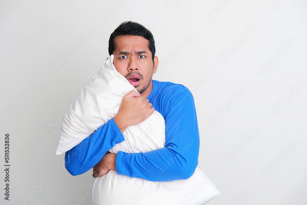 Adult Asian man hugging a pillow while showing scared face expression Adult Asian man hugging a pillow while showing scared face expression