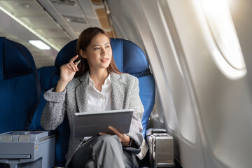 Successful beautiful young asian business woman sits in airplane cabinplane and works on digital tablet with stylus. Flying at first class.