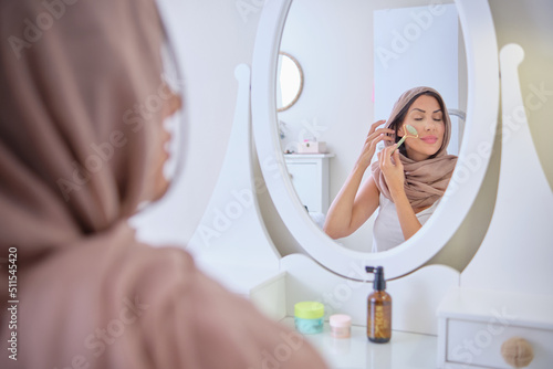 young Muslim woman wearing hijab headscarf putting on makeup. Muslim woman applying make-up against blue colour background. Young Muslim woman with mirror and makeup brush and jade 