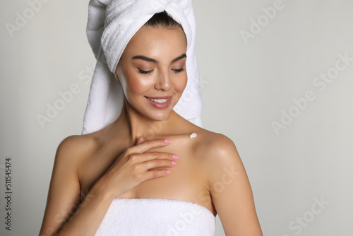Beautiful young woman with towels applying cream on shoulder against light background