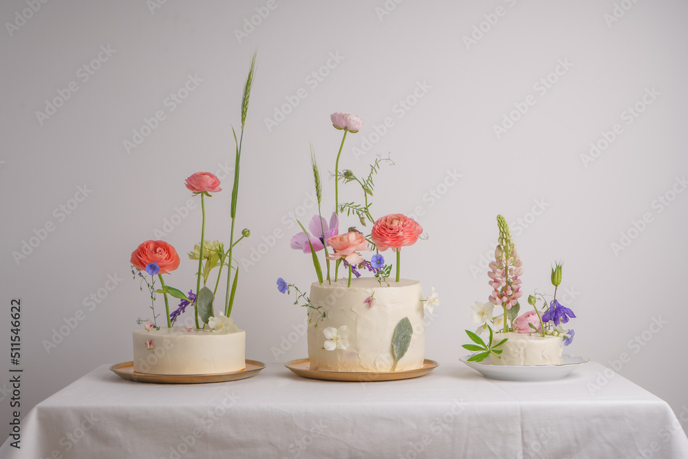 Three white beautiful wedding cake with cream and colored fresh flowers on a white table with a tablecloth and a white background