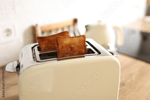 Toasted bread and toaster for breakfast. Modern white toaster and roasted bread slices toasts inside on wooden table in kitchen.