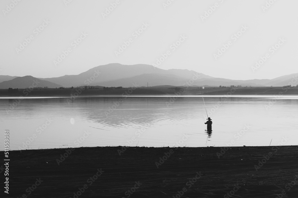 A man is fishing in the lake at sunset. Wetland and mountain landscape. Black and white.