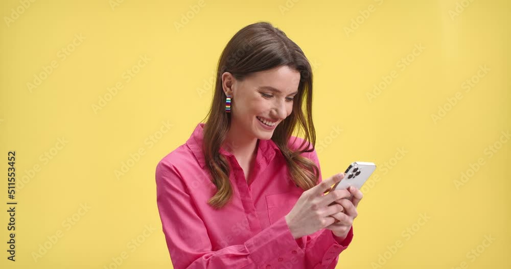 Close up portrait of happy young pretty woman smiling while typing on smartphone on yellow background. Cheerful beautiful female in pink blouse on yellow wall using mobile phone looking at screen