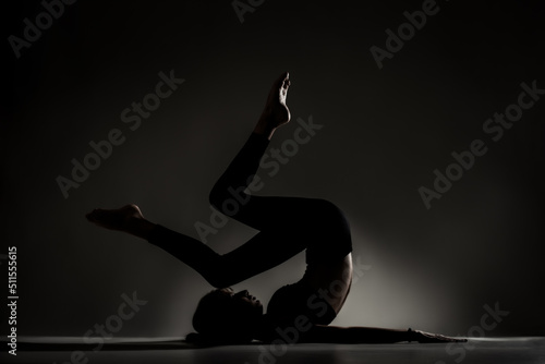 Fit ballerina girl lying and stretching practicing yoga poses. Side lit silhouette against dark background.. .