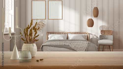 Wooden table, desk or shelf close up with ceramic and glass vases with dry plants, straws over blurred view of wooden bedroom with double bed, modern interior design concept