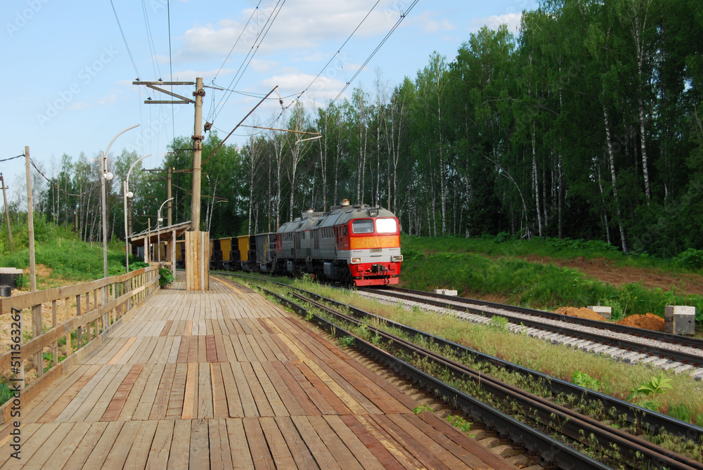 A freight train rides past a wooden country station. Transportation of goods by rail. The freight train transports goods. Cargo transportation by rail.