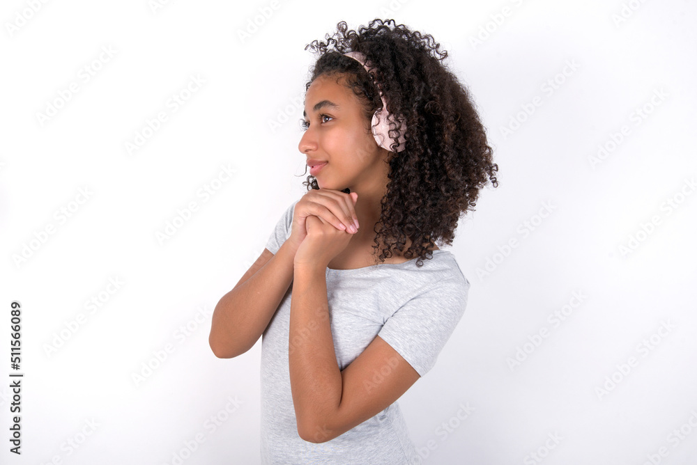 Young beautiful girl with afro hairstyle wearing grey t-shirt over white wall wears stereo headphones listening to music concentrated and looking aside with interest. Young beautiful girl with afro hairstyle wearing grey t-shirt over white wall wears stereo headphones listening to music concentrated and looking aside with interest.