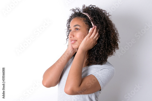 Young beautiful girl with afro hairstyle wearing grey t-shirt over white wall wears stereo headphones listens music concentrated aside. People hobby lifestyle concept