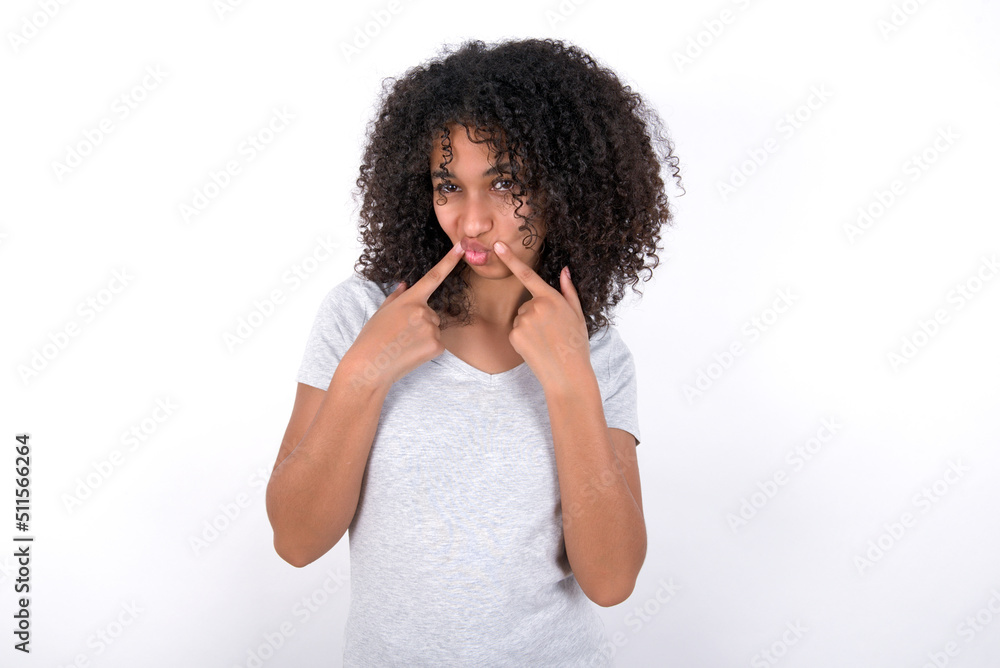 Young beautiful girl with afro hairstyle wearing gray t-shirt over white background crosses eyes and makes fish lips funny grimace Young beautiful girl with afro hairstyle wearing gray t-shirt over white background crosses eyes and makes fish lips funny grimace