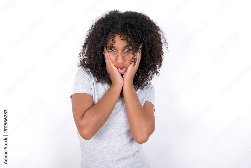 Young beautiful girl with afro hairstyle wearing gray t-shirt over white background keeps hands on cheeks has bored displeased expression. Stressed hopeless model