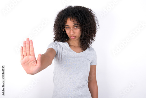 Young beautiful girl with afro hairstyle wearing gray t-shirt over white background shows stop sign prohibition symbol keeps palm forward to camera with strict expression